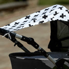 Ein grauer Kinderwagen wird an einem sonnigen Tag im Freien mit unscharfem Hintergrund von dem Sonnensegel schwarze Pluszeichen von KraftKids aus 100 % Baumwolle beschattet, das mit kräftigen schwarzen Pluszeichen versehen ist.