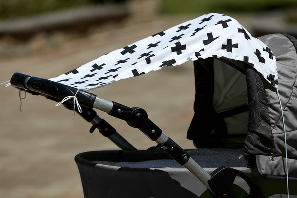 Ein grauer Kinderwagen wird an einem sonnigen Tag im Freien mit unscharfem Hintergrund von dem Sonnensegel schwarze Pluszeichen von KraftKids aus 100 % Baumwolle beschattet, das mit kräftigen schwarzen Pluszeichen versehen ist.