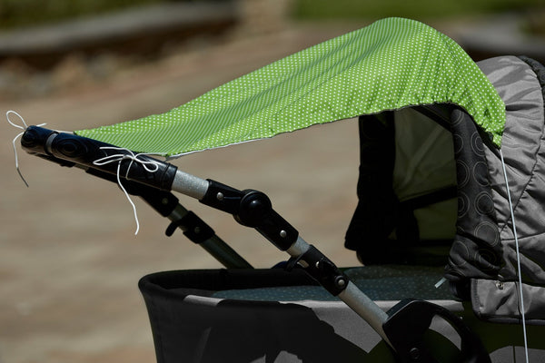 Nahaufnahme eines Kinderwagens im Freien an einem sonnigen Tag, mit dem KraftKids Sonnensegel weiße Punkte auf Grün, das mit weißen Schnüren für zusätzliche Sicherheit gesichert ist.