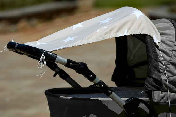 Nahaufnahme eines grauen Kinderwagens mit einem KraftKids Sonnensegel große weiße Sterne auf beigem Baumwollverdeck. Die Kanten haben ausgefranste Fäden, und der Hintergrund im Freien ist unscharf.