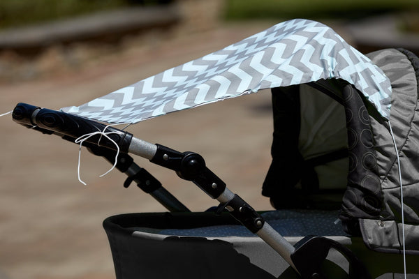 An einem grauen Kinderwagen ist der KraftKids Sonnensegel Chevron hellgrau und mint an den Griffen befestigt und bietet zusätzlichen Sitzschutz. Der Hintergrund ist im Freien und unscharf.
