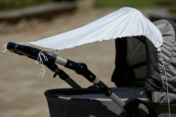 Eine Nahaufnahme zeigt einen Kinderwagen, über dessen Verdeck und Griffe das KraftKids Sonnensegel Karo grau drapiert ist, das den Kinderwagen effektiv vor der Sonne schützt. Der sanft verschwommene Hintergrund hebt den beschatteten Kinderwagen hervor.