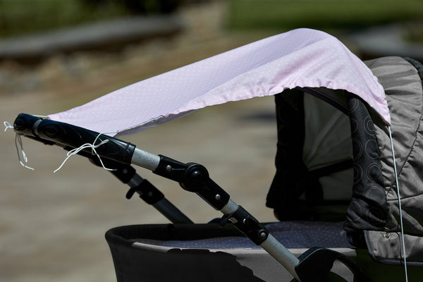 Eine Nahaufnahme eines KraftKids Sonnensegel weiße Punkte auf Rosa zeigt sein hellrosa, gepunktetes Verdeck, das lose an einem grau-schwarzen Kinderwagengestell befestigt ist, wobei leichte Ausfransungen sichtbar sind, vor einem unscharfen Hintergrund im Freien.