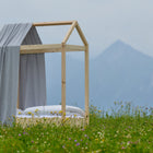 Das KraftKids Bettbaldachin Musselin grau Pusteblumen bringt kreative Kinderzimmerdekoration, mit grauem Musselinhimmel und weißer Bettwäsche, wunderschön eingebettet in eine Wildblumenwiese mit Bergblick unter einem wolkigen Himmel.