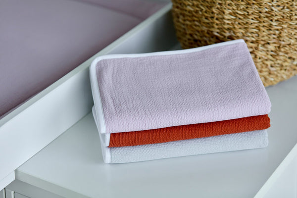 Drei pastellfarbene Handtücher in Rosa, Orange und Grau werden von einem geflochtenen Korb und der KraftKids Wickelunterlage Doppelkrepp Rosa gestapelt - ideal für die Bedürfnisse Ihres Babys beim Wickeln.
