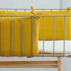 Eine Nahaufnahme eines modernen Babybettes mit einem Metallrahmen, Holzbeinen und der KraftKids Bettumrandung Nestchen Doppelkrepp Gelb Senf an den Seiten befestigt. Die Matratze ist mit einem weißen Laken vor einem schlichten Hintergrund bedeckt - die perfekte Babyausstattung.