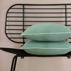 Zwei KraftKids Kissenbezug Doppelkrepp Grün Jade mit weißer Paspel sind auf einem modernen schwarzen Drahtstuhl vor einer beigen Wand gestapelt.