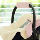 Eine Nahaufnahme der KraftKids Decke für Babyschale Winter Doppelkrepp Rosa in zarten Creme- und Rosatönen mit Gurtschlitzen, die in einem Innenraum in der Nähe eines Fensters mit Jalousien und einem verschwommenen grünen Blick nach draußen platziert ist.