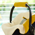 Eine Nahaufnahme der KraftKids Decke für Babyschale Winter Doppelkrepp Gelb Senf mit Gurtschlitzen, dargestellt in einer Babyschale mit gelbem Griff und weißer Polsterung, vor einem unscharfen Hintergrund mit Jalousien und Grünzeug.