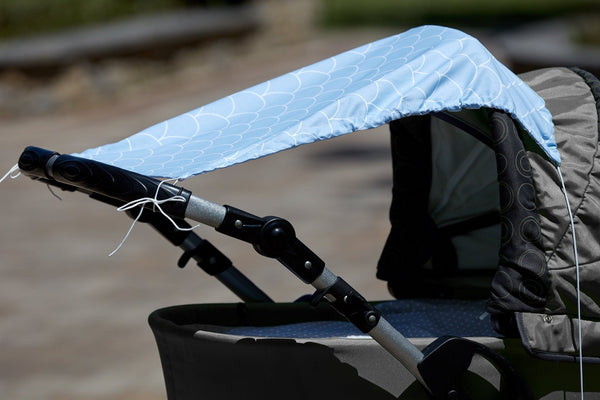 Eine Nahaufnahme eines Kinderwagens mit dem KraftKids Sonnensegel weiße Halbkreise auf Pastellblau. Der Sonnenschirm aus Baumwolle schützt Ihr Kind im Freien, während ein sanft verschwommener Hintergrund sein stilvolles weißes Halbkreismuster hervorhebt.