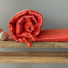 Eine aufgerollte KraftKids Bettumrandung Nestchen Cord Breitcord in Reinorange mit weißer Paspel und Bändern liegt auf einer rustikalen Holzbank, neben zwei glatten Holzkugeln, vor einem schlichten grauen Hintergrund.