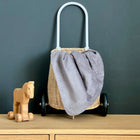 Ein Weidenkorb mit Rädern und einem geschwungenen Griff steht auf einer Holzfläche. Darüber drapiert ist die KraftKids Babydecke Cord Breitcord Grau. Neben dem Korb steht ein kleines Holzspielzeugpferd auf Rädern, alles vor einer schlichten dunklen Wand.