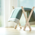 Ein heller Holzstubenwagen mit einer weichen grün-weißen KraftKids-Decke steht an einem großen Fenster. In der Nähe steht ein geflochtener Korb mit rosa Bommeln und eine kuschelige KraftKids Krabbeldecke Musselin mint, die mit Polyestervlies gefüllt ist.