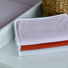 Ein Stapel von drei gefalteten Handtüchern in Pastellrosa, Orange und Hellgrau liegt auf einer weißen Fläche neben einem geflochtenen Korb, auf dem die KraftKids Wickelunterlage Doppelkrepp Rosa fein säuberlich platziert ist.