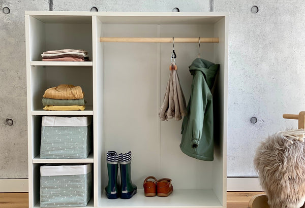 Der KraftKids Kleiderschrank für Kinder nach Montessori in Weiß bietet Regale mit gefalteter Kleidung, gemusterte Behälter, eine Stange mit einem Schal und einer grünen Jacke sowie Regenstiefel und Sandalen auf dem unteren Regal vor einer Betonwand.