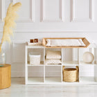 Eine moderne Wickelkommode zeigt ein weißes Regal mit gefalteten Decken, einem Korb und der kuscheligen KraftKids Wickelauflage Musselin Zweige braun. Ein Holzhocker mit einer Glasvase mit Pampasgras steht vor der weiß getäfelten Wand.