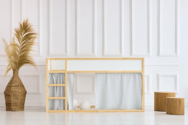 Ein minimalistisches Kinder-Etagenbett mit Holzrahmen und KraftKids Hochbettvorhänge Set 2+1 Musselin grau Pusteblumen, passend zum Kura Bett, in einem hellen Zimmer mit weiß getäfelten Wänden, dazu eine große Korbvase mit Pampasgras und zwei Holzhocker.